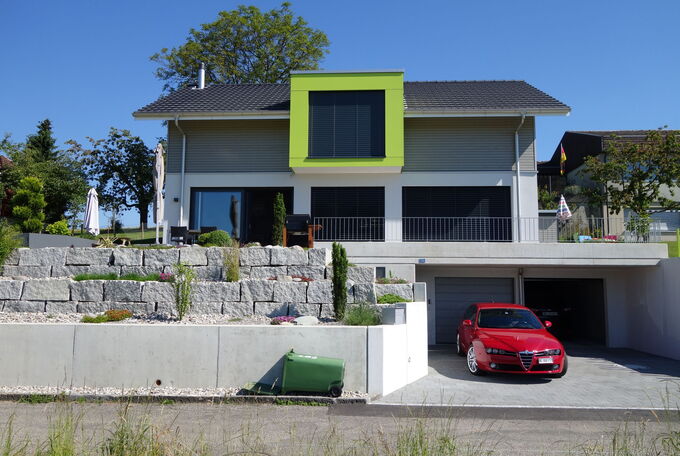 Frontansicht EFH, das Haus steht auf der Garage an erhöhter Lage, eine grüne Lukarne im OG sticht ins Auge