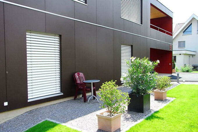 Rechteckiges Haus mit integriertem gedecktem Sitzplatz im OG, dunkelbraune mit roten Details kombinierte Vollkernplattenfassade