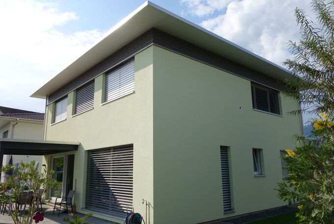 Hausansicht von der Gartenseite her, die silbernen Lamellenstoren bei den Fenstern sind geschlossen, harmonieren schön mit der Holzschlung unter dem Dach