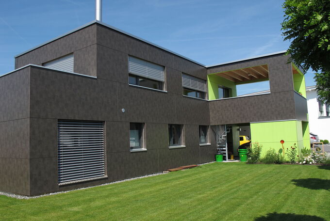 Haus mit Flachdach, dunkle Fassade kombiniert mit apfelgrünen Details und zwei grossen Terrassen im Obergeschoss, eine davon gedeckt