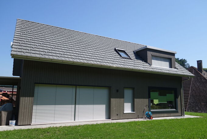 Hausfassade mit dunkler Holzschalung, grosser Fensterfront zum Garten und Lukarne im Obergeschoss